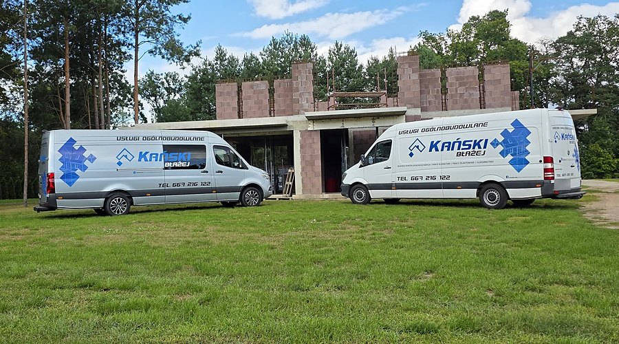 Dwa firmowe busy z logo Kański przed budynkiem w stanie surowym